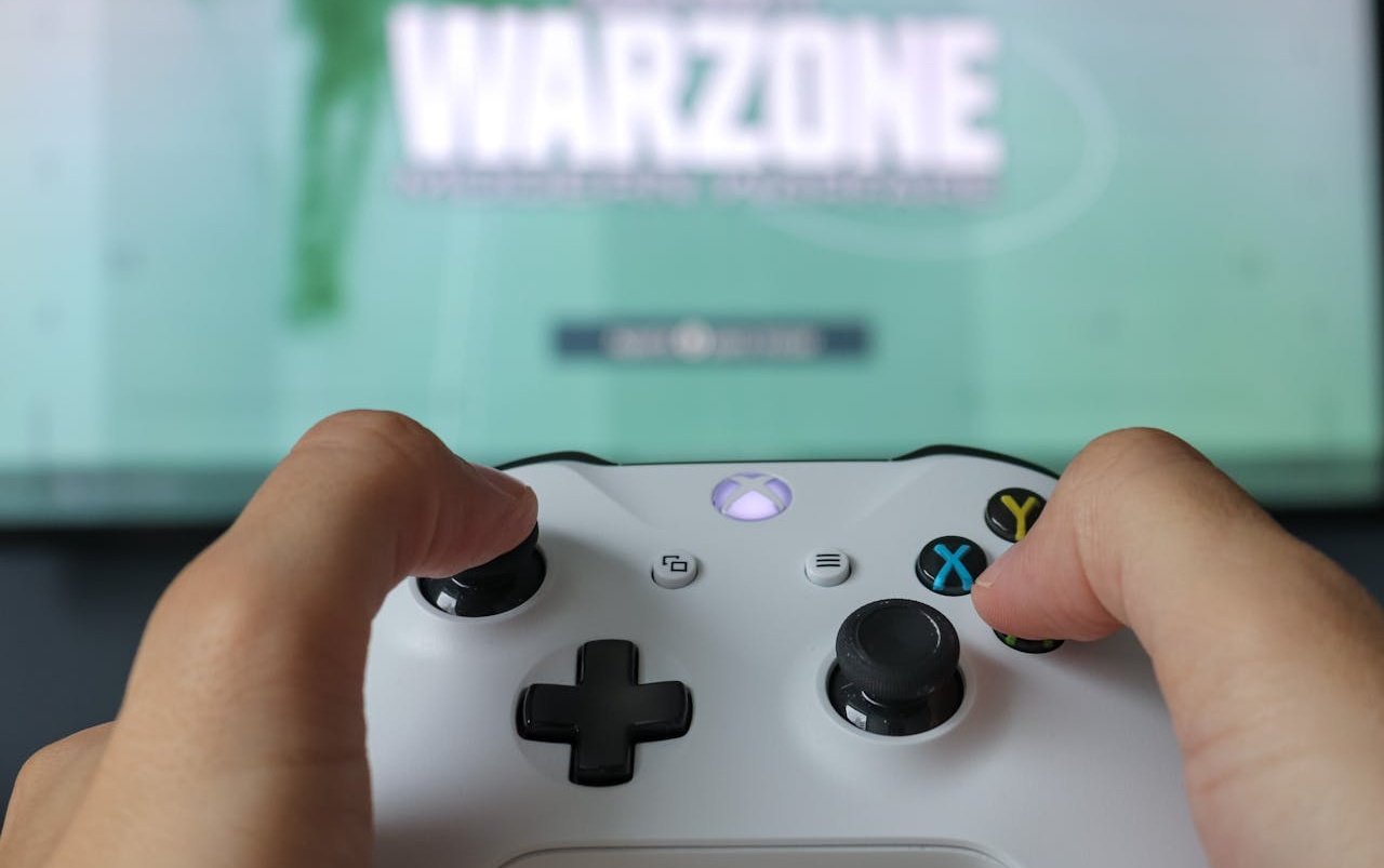 person-holding-a-white-xbox-controller-warzone-game-title-on-a-blurred-tv-screen-in-the-background - Online Tech Tips A person holding a white Xbox controller with the game title "WARZONE" displayed on a blurred TV screen in the background