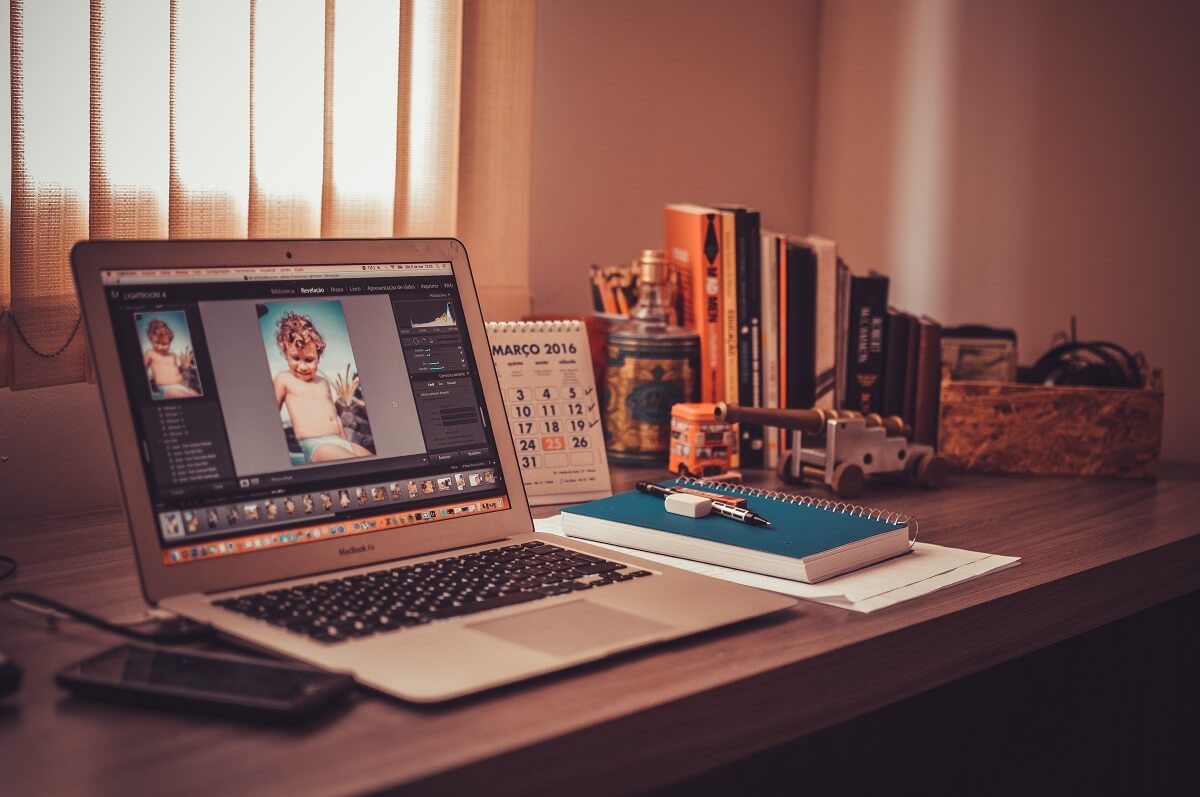 A laptop displays photo editing software, showcasing a toddler's picture; it rests on a desk cluttered with books, a calendar reading "MARÇO 2016", and other items