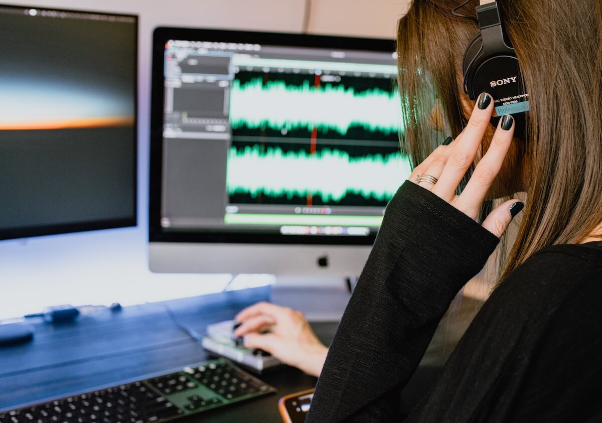 A woman adjusts headphones while working at a computer displaying audio waveforms