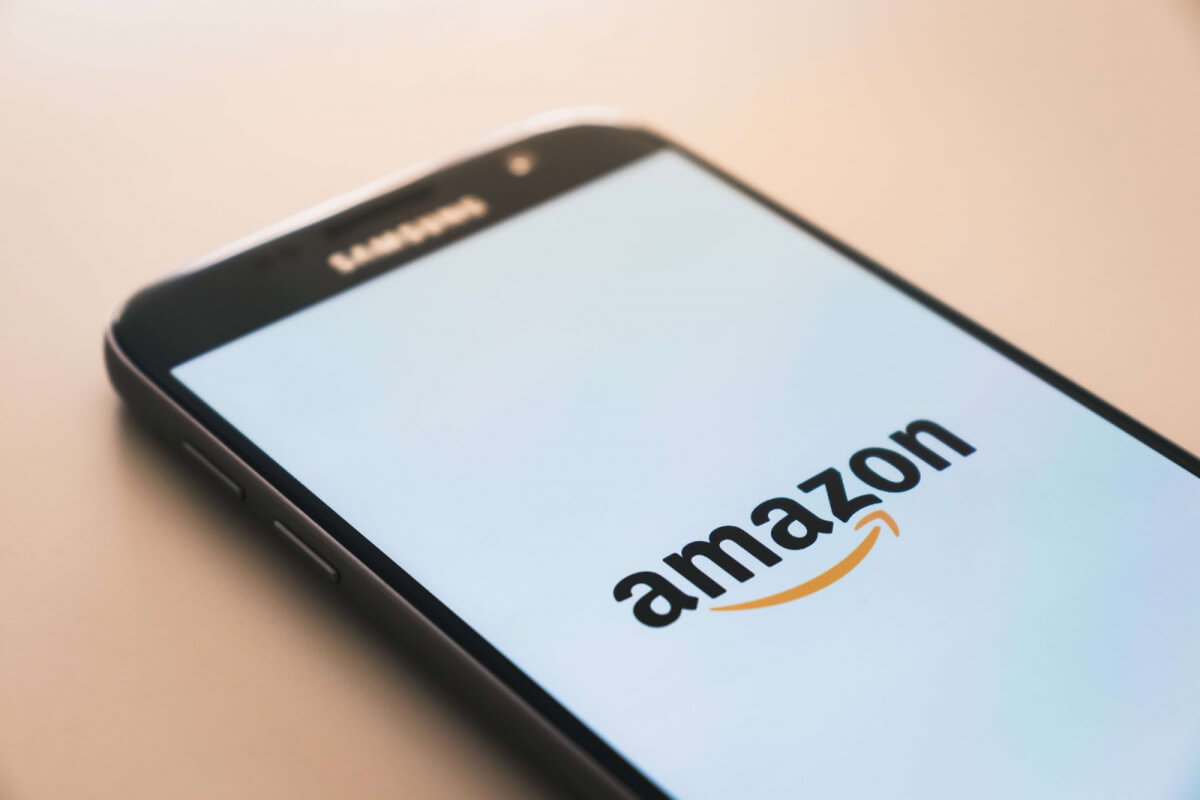 A smartphone displays the Amazon logo on its screen; it rests on a light beige surface