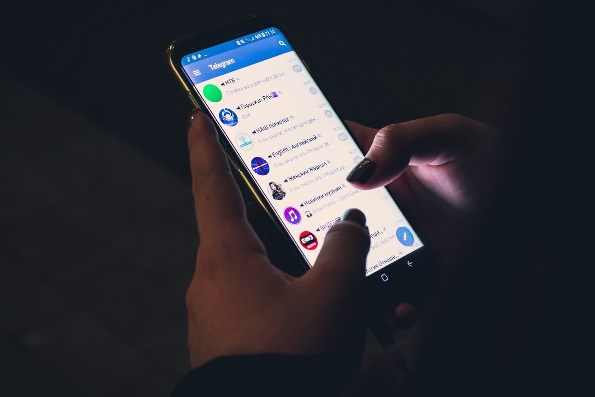 A person's hands interact with a smartphone displaying the Telegram app, actively scrolling through a list of channels in a dimly lit environment