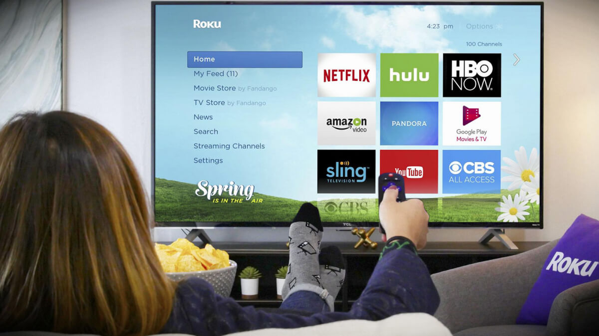 A woman uses a Roku remote to navigate a streaming service menu on a large TV screen displaying various app icons, while relaxing on a couch with snacks